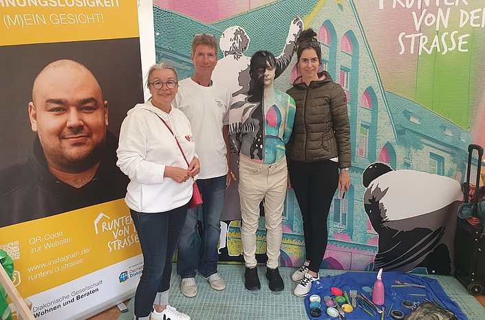 Vier Personen stehen neben einer bunt bemalten Schaufensterpuppe vor einer Graffiti‑Wand und einem Informationsbanner.
