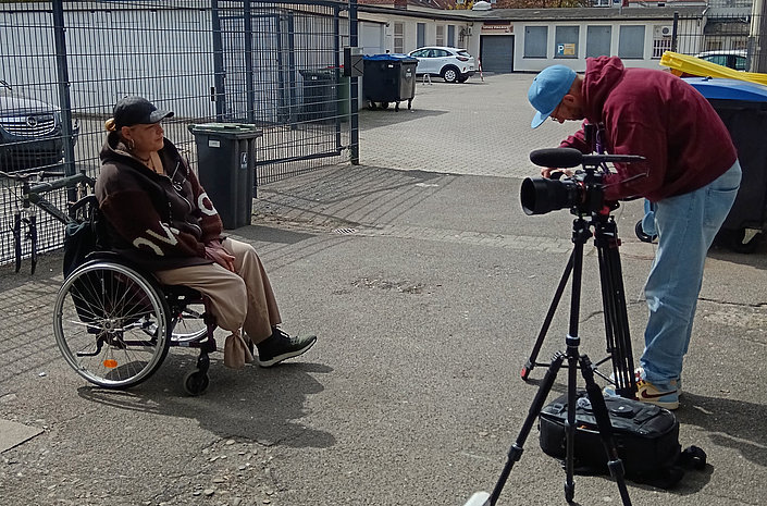 Alternativtext (≤155 Zeichen) Person im Rollstuhl sitzt auf einem Hof und wird von einem Kameramann mit Stativ gefilmt; Zaun, Mülltonnen und Gebäude im Hintergrund.