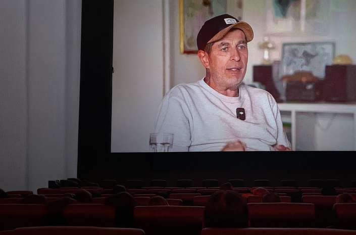 Dunkler Kinosaal mit großer Leinwand, darin ein Mann im Interview; im Publikum ist ein leuchtendes Handydisplay sichtbar.
